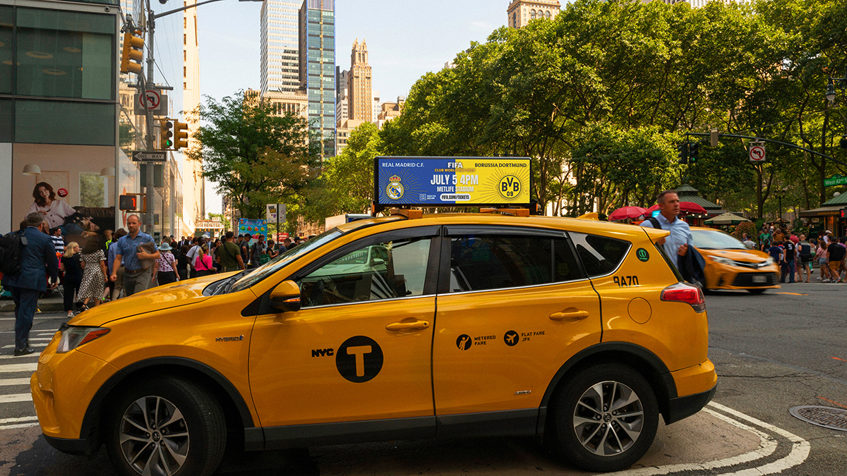 Yellow NYC taxi with a blue digital LED top ad promoting a FIFA match at MetLife Stadium, driving on a Manhattan street